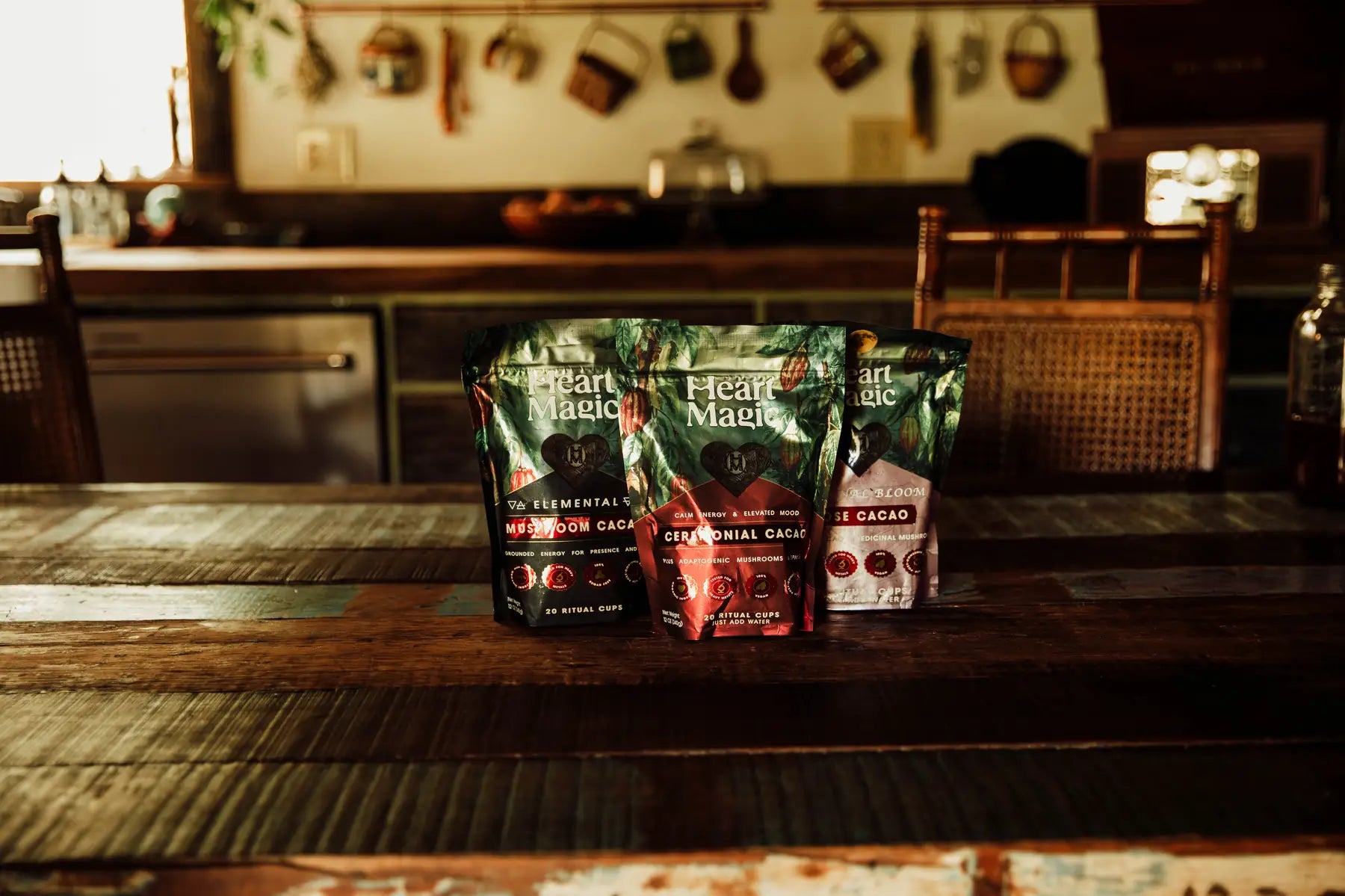 Three bags of Heart Magic cacao hearts, Elemental, Ceremonial and Rose, on a worn wooden kitchen table in warm evening light