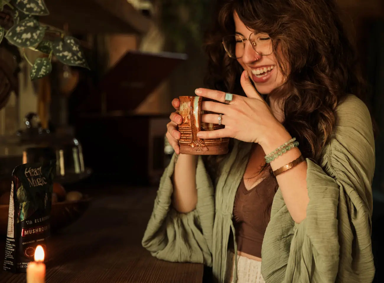 A woman laughs while holding a mug of cacao in both hands beside a candle and a bag of Elemental