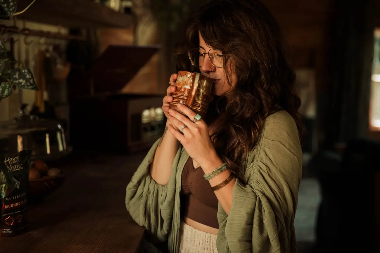 A woman with eyes closed sips cacao from a speckled ceramic mug in a warm, dim kitchen